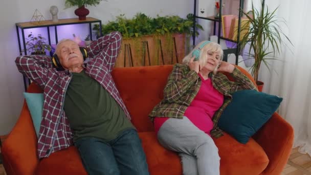 Счастливые пожилые пожилые семьи пара бабушка и дедушка мужчина женщина носит наушники прослушивания музыки расслабляющий сидя на диване дома в выходные. Зрелая бабушка дедушка отдыхает вместе на диване. - Кадры, видео