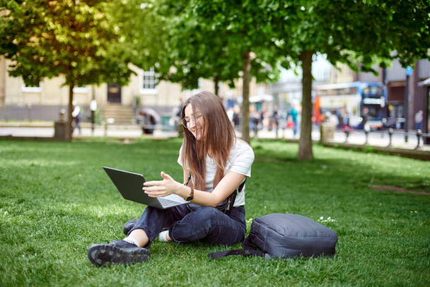 A young girl works with a laptop in the fresh air in the park, sitting on the lawn. The concept of remote work. Work as a freelancer. The girl takes courses on a laptop and smiles. - Foto, immagini