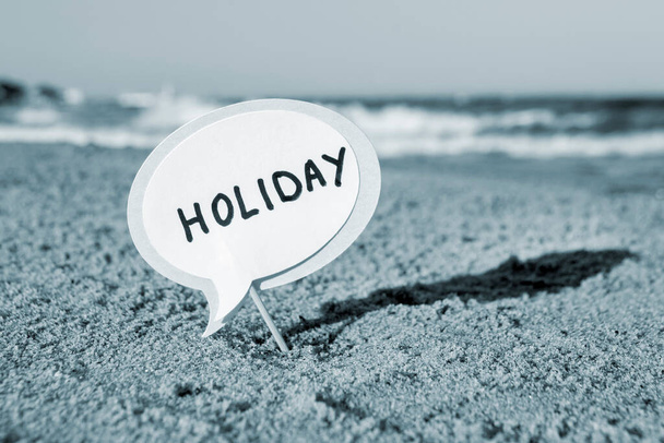 Small stick with paper speech bubble with word Holiday stands on sandy beach, sea water and sea waves with white foam close-up. Concept of travel tourism vacation holiday relax rest summer. Blue color - Photo, Image
