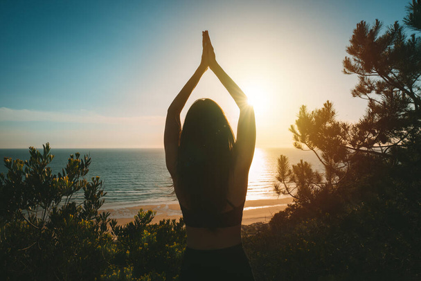 Silhouette of young healthy woman practicing yoga with ocean view at sunset. Rear view of woman standing with hands joined over head in tree pose. High quality photo - Photo, image