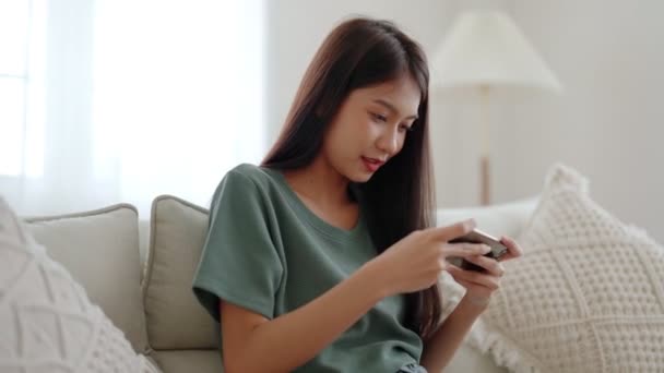Szczęśliwa młoda azjatycka kobieta zrelaksować się na wygodnej kanapie w domu, gry online na smartfonie, uśmiechnięta dziewczyna korzystać z telefonu komórkowego czat, przeglądać bezprzewodowy internet na gadżet, zakupy online z domu - Materiał filmowy, wideo