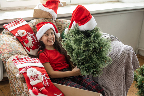 Foto von niedlichen fröhlichen kleinen Mädchen macht einen künstlichen Weihnachtsbaum tragen Weihnachtsmütze drinnen. - Foto, Bild
