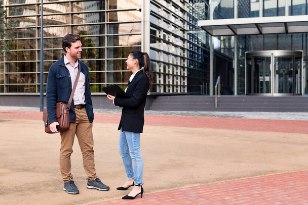 caucasian man and asiatic woman smiling and talking about business in front of an office building, concept of coworkers and lifestyle, copy space for text - Fotoğraf, Görsel