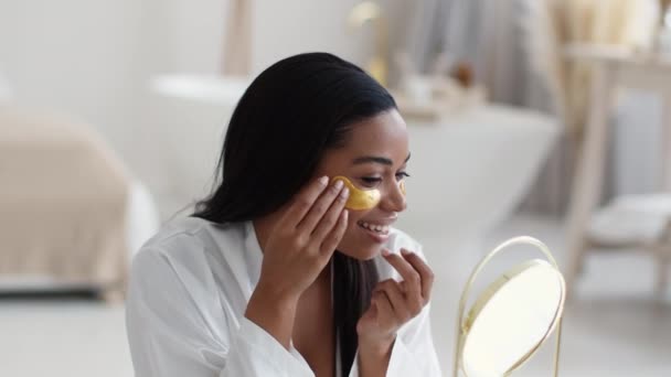 Huomenta, kaunotar. Nuori onnellinen afrikkalainen amerikkalainen nainen kylpytakissa laittaa kultaiset kollageenilaastarit silmien alle, päästä eroon pusseista, hymyillen peilille - Materiaali, video
