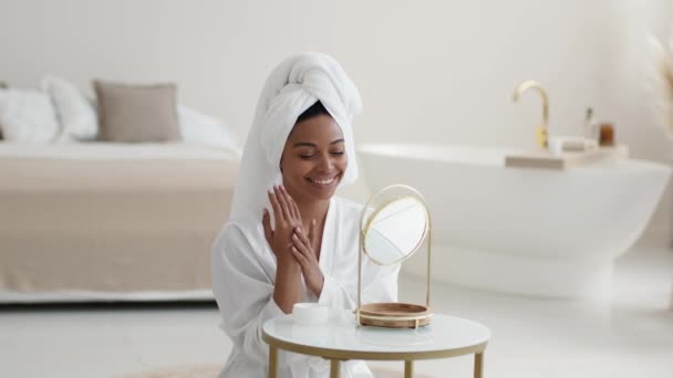 Jonge mooie Afrikaanse Amerikaanse dame met handdoek op het hoofd aanbrengen hydraterende crème op handen, verwennen lichaam na douche, kijken naar spiegel op badkamer, vrije ruimte - Video