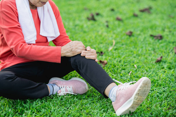Femme asiatique âgée avec douleur à la cheville au genou tout en courant dans le parc. Senior asiatique femme assise sur le sol et tenant genou douloureux. - Photo, image