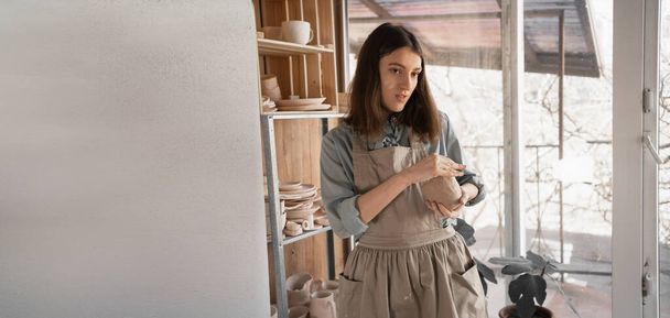 Egy fiatal kézműves nő arra koncentrált, hogy nyers agyagból faragjon egy kancsót egy kreatív stúdióban tartott kerámiaórán vagy műhelyben. Egy fiatal kerámiaművész kerámiával foglalkozik.. - Fotó, kép