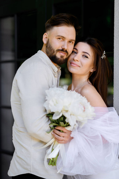 Gyönyörű kaukázusi friss házaspár fehér ruhában, fehér bazsarózsa csokorral, ölelkezve és csodálva a ház hátterét - Fotó, kép
