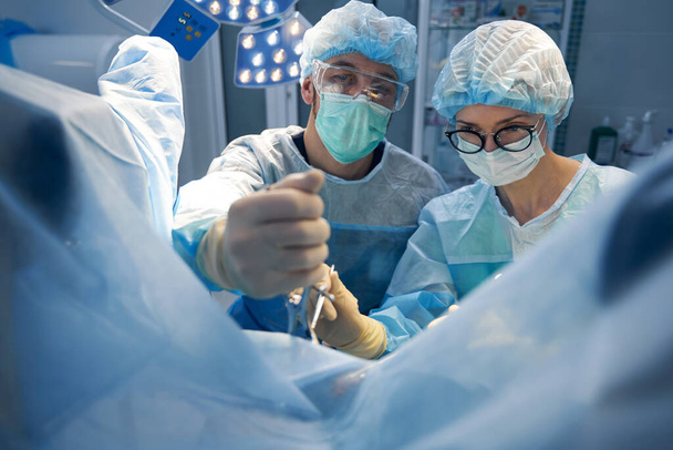 Male medic holding tissue in incision with locking forceps while nurse using surgical scissors - Foto, Imagem
