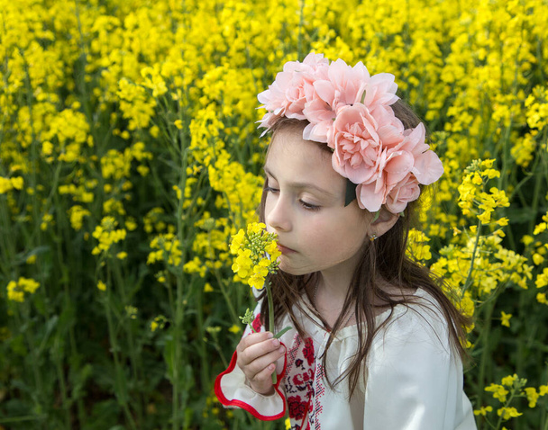 Portrait of a cute girl 7 years old in a flower wreath on her head among a yellow blooming rapeseed field, smelling a flower. Children for Peace. Support Ukraine. Stop the war. Pride to be Ukrainian - Photo, Image
