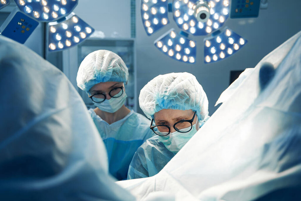 Пожилая женщина хирург и медсестра в очках, проводящая лапароскопические процедуры на пациенте - Фото, изображение