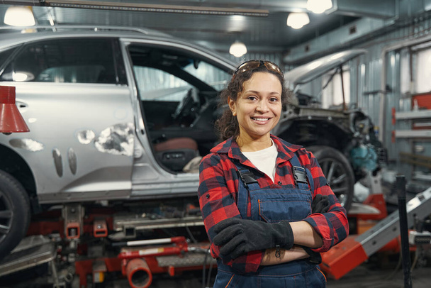 Joyful female worker keeping arms crossed and smiling while standing near vehicle in repair service station - Fotó, kép