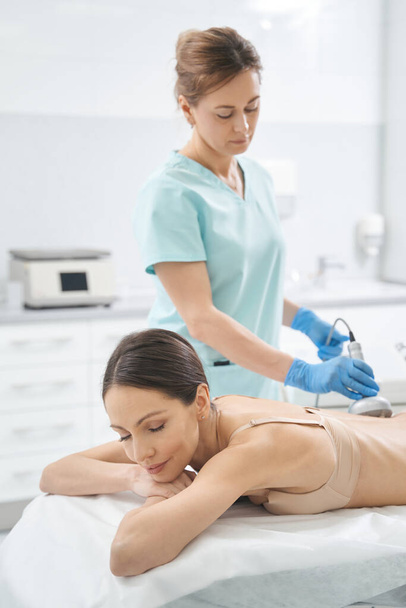 Kobieta na łóżku w staniku otrzymuje RF-terapię pleców od kosmetyczki - Zdjęcie, obraz