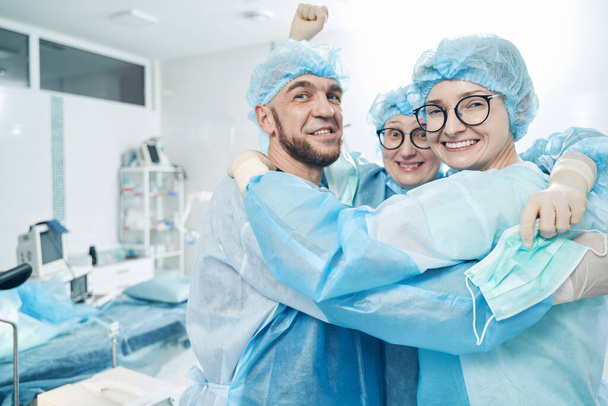 笑顔でカメラを探し、手術室で空気中で拳を上げる3人の医療従事者 - 写真・画像