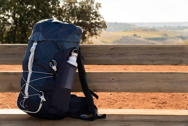 Пішохідний рюкзак з пляшкою на дерев'яній лавці в сільській місцевості з тлом гір - спортивно-оздоровча концепція
 - Фото, зображення