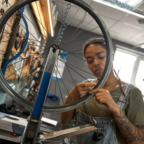 Young african american female cycling repairman checking bicycle wheel spoke with bike spoke key in modern workshop. Bike service, repair and upgrade. Garage interior with tools and equipment - 写真・画像