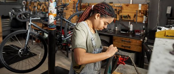Female cycling master fixing bike tube in modern bicycle workshop. Young concentrated black girl. Bike service, repair and upgrade. Garage interior with tools and equipment. Daytime - Photo, image