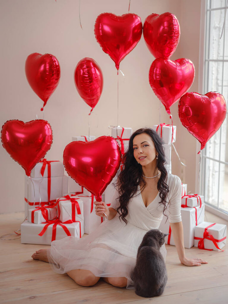 Hermosa joven feliz en vestido blanco y gato gris. fiesta de vacaciones. Modelo alegre posando con globos en forma de corazón rojo, divirtiéndose, celebrando el Día de San Valentín. Símbolo del amor - Foto, imagen
