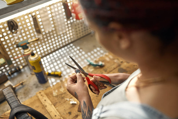 Partial image of black female cycling master cutting bicycle patch with scissors for glueing on bike tube in blurred modern workshop. Woman with tattoo. Bike service, repair and upgrade - Fotó, kép