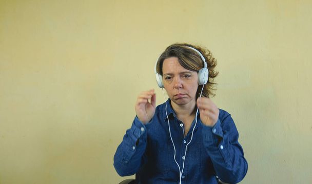 デニムのシャツを着た40歳の女性は、大きな有線の白いヘッドフォンで音楽を聞いています。女の肖像画 - 写真・画像