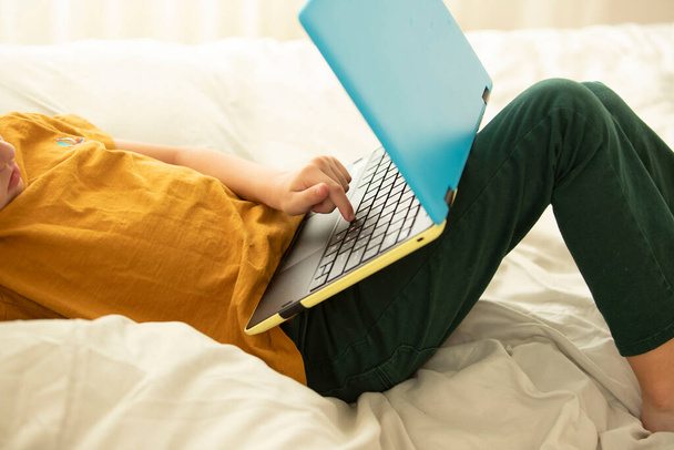 Дитина лежить на ліжку з ноутбуком в жовто-блакитних кольорах, друкуючи пальцем на клавіатурі. Частина тіла. Концепція віртуальних ігор, онлайн-освіта, комфортне навчання, інтернет-зв'язок
.  - Фото, зображення
