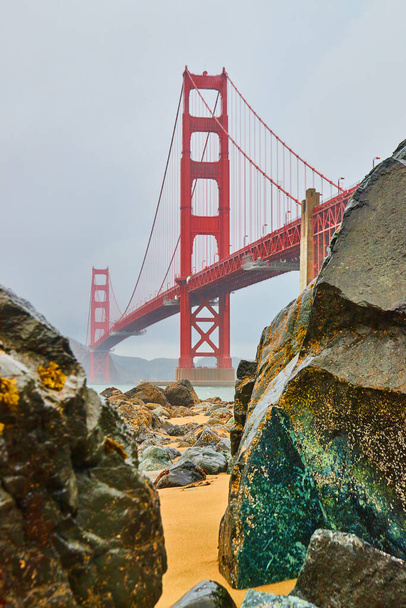 Image du Golden Gate Bridge à travers un trou dans les roches sur une plage de sable fin pendant la matinée brumeuse - Photo, image