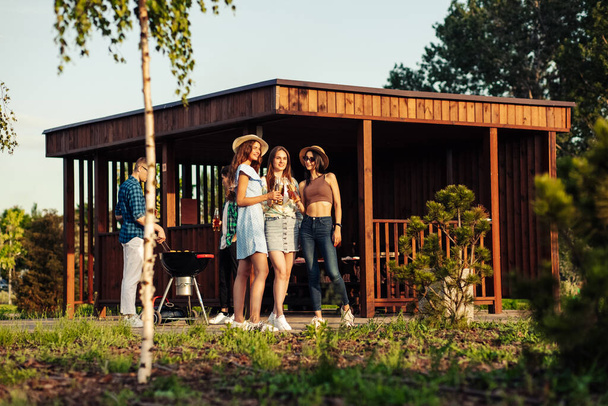 Three beautiful girls are drinking drinks in glass bottles and talking, in the background a group of friends having fun grilling meat enjoying barbecue, preparing and eating barbecue dinner outdoors. Men and women stand around the grill and chat - Foto, Imagen