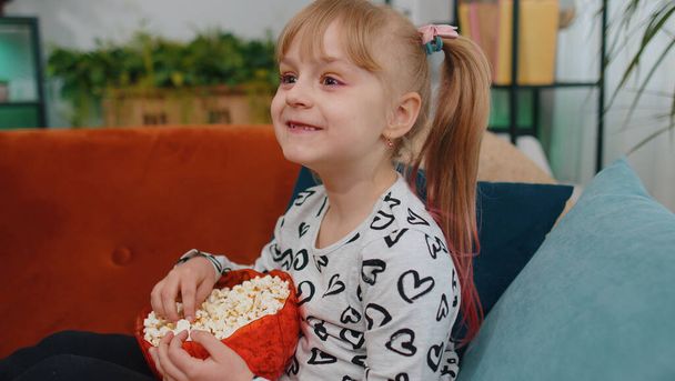 Young funny little teenager children girl watching comedy video show content film on tv social media, eating popcorn on sofa couch at home alone. Small cute school toddler child kid. Relaxation time - Photo, Image