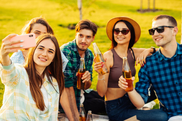 Happy friends have a picnic in the city park outdoors. Young people drink drinks and eat pizza and laugh in the street while sitting on a ravine. Friends have fun talking and taking selfie on mobile phone - Zdjęcie, obraz