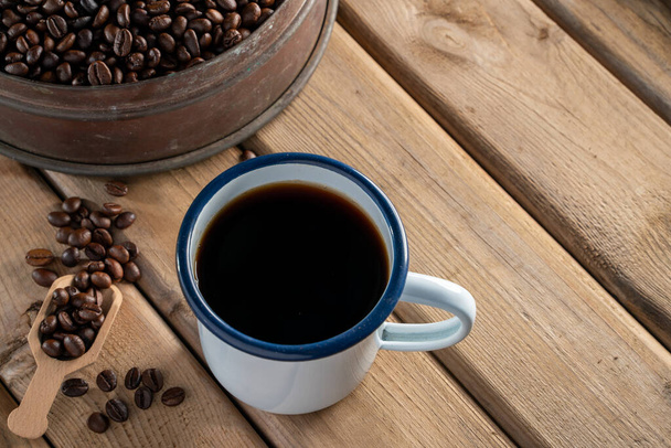 bílý smaltovaný hrnek na kávu a tmavá kávová zrna na staré dřevěné podlaze. soft focus.shallow efekt zaostření. - Fotografie, Obrázek