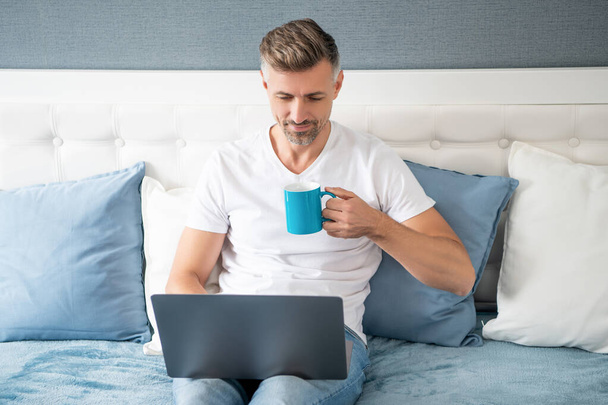 busy mature man using computer in bed with coffee. - Foto, Imagen