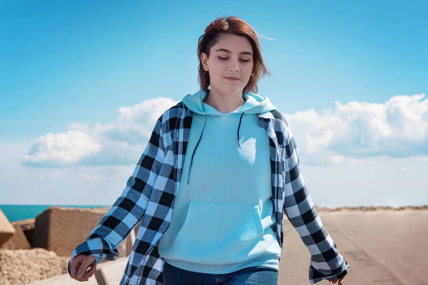 Short haired teenager in light blue hoodie and plaid shirt. Teen girl walking outdoor on the road and looking down, sunny day and blue sky. Clothing mockup, freedom adolescence concept - Photo, Image
