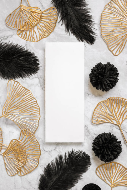 Blank card on marble table near black feathers and golden leaves with wedding rings top view. Menu or Invitation card mockup. Elegant femminine flat lay with vertical paper card  - Foto, Imagen