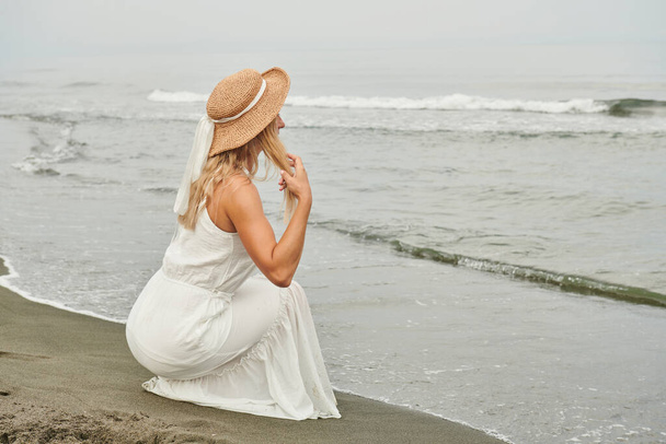 Ein Mädchen in weißem Kleid und Hut sitzt am Strand. Hochwertiges Foto - Foto, Bild