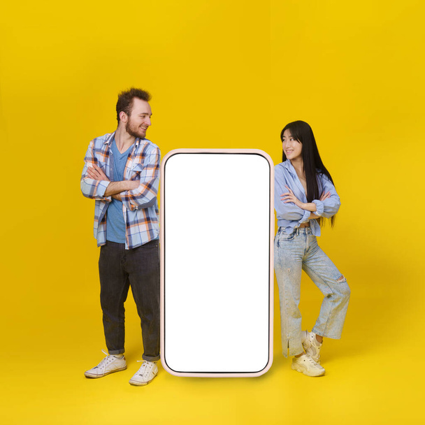 Asian girl and caucasian guy standing leaned on huge smartphone with white blank screen, mobile app advertisement and smile looking at each other isolated on yellow background. Product placement. - Valokuva, kuva