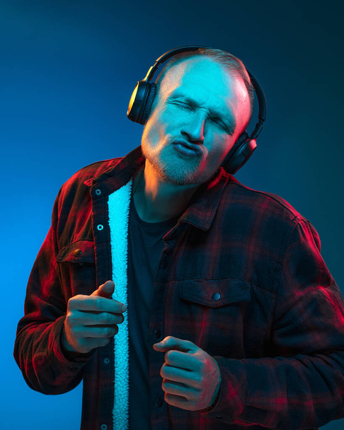 Portrait of emotional man on blue studio background in red cyan neon, multicoloured. Listening to music with headphones with pleasure and enjoyment. Flyer Poster Banner Advertising - Zdjęcie, obraz
