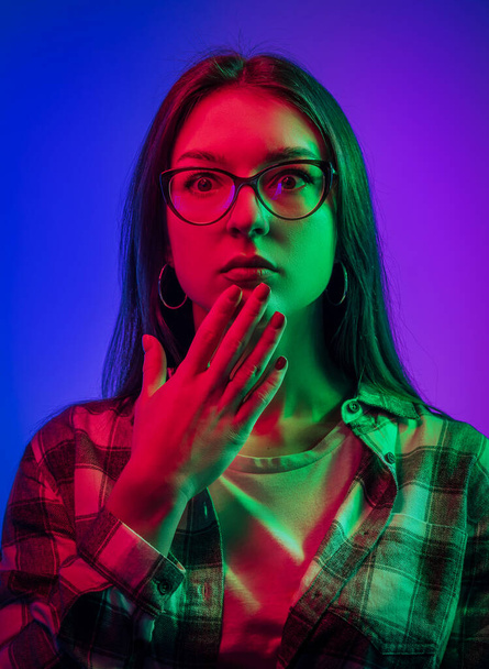 Portraits of young emotional woman on multicoloured background in neon lights. Looks with unexpected surprise and holds his hand to his mouth. Place for text. Flyer Poster Banner - Fotoğraf, Görsel