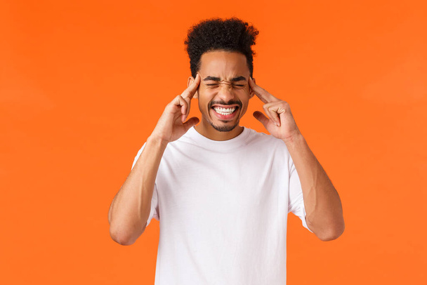 Distressed and tensed african-american male in white t-shirt, clench teeth, close eyes and touch temples, feeling migraine or headache, trying calm down and think straight, orange background. - Foto, imagen