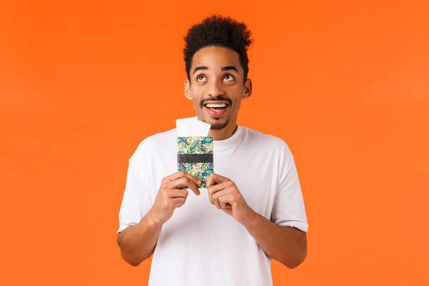 Dreamy cheerful, smiling african-american guy with moustache, afro haircut, holding passport with flight tickets, looking up grinning joyfully, picturing himself on luxury resort, orange background. - Photo, Image