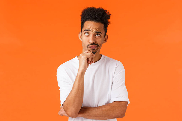 Need to think about it. Hesitant and unsure, thoughtful african-american moustached hipster guy, smirk doubtful, touch chin and looking up thinking, standing indecisive over orange background. - Photo, Image
