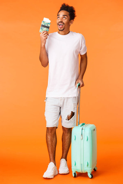 Plan vertical complet rêveur, excité homme afro-américain en vacances, tenant des bagages, valise de transport et passeport avec billets d'avion, voyage parfait d'imagerie, voyage impressionnant, fond orange
. - Photo, image
