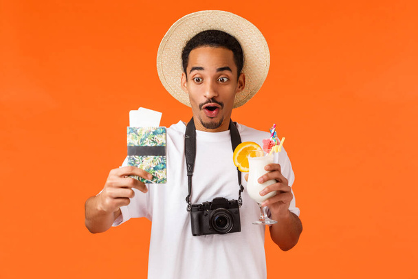 Divertido cara feliz afro-americano finalmente em férias, indo para o exterior, segurando coquetel e passaporte com dois bilhetes, olhando espantado e alegre, sorrindo, de pé sobre fundo laranja
. - Foto, Imagem