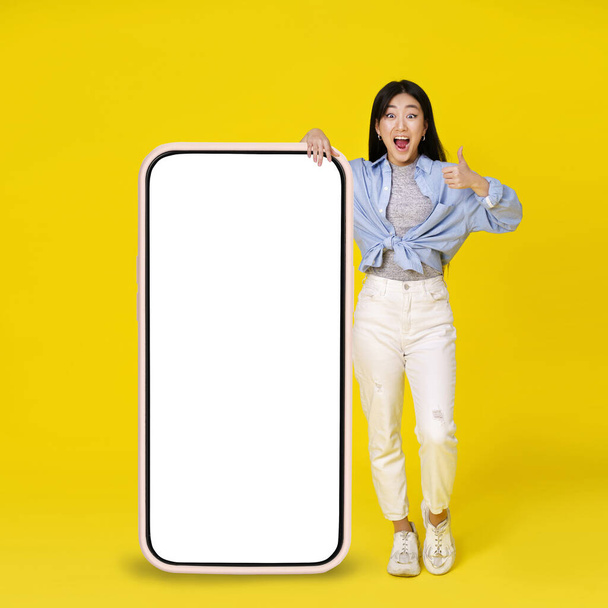 Excited asian girl gesturing thumb up leaned on giant, huge smartphone with white, blank screen standing leaned on wearing casual isolated on yellow background. Free space mock up. - Fotó, kép