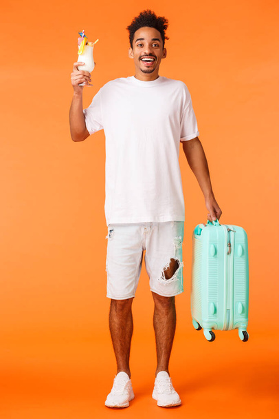 フル長さの垂直ショット幸せなアフリカ系アメリカ人の男の休暇では、旅行として荷物を保持し、ホテルでハイフレンドと言って、カクテルを飲み、喜んで笑みを浮かべて。オレンジの背景に立って. - 写真・画像
