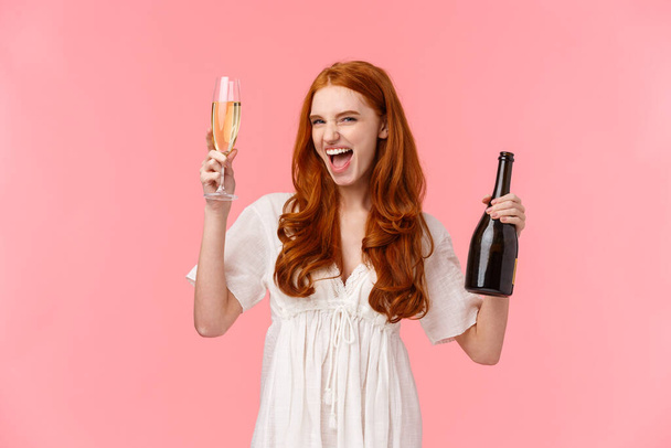 Podekscytowana i beztroska pijana ruda biała dziewczyna dobrze się bawiąca, szalejąca i wolna na niesamowitej imprezie, podnosząca szklankę i butelkę szampana, uśmiechnięta pyskata, stojąca na różowym tle. - Zdjęcie, obraz