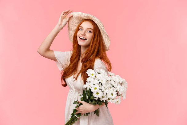 Κορίτσι που απολαμβάνει την ανοιξιάτικη μέρα κάτω από τον ήλιο, stralling αγορά μπουκέτο για τη φίλη, κρατώντας λευκά λουλούδια και χαμογελαστή κάμερα, αγγίζοντας cheeky καπέλο και υπέροχη ατενίζοντας, στέκεται ροζ φόντο. - Φωτογραφία, εικόνα