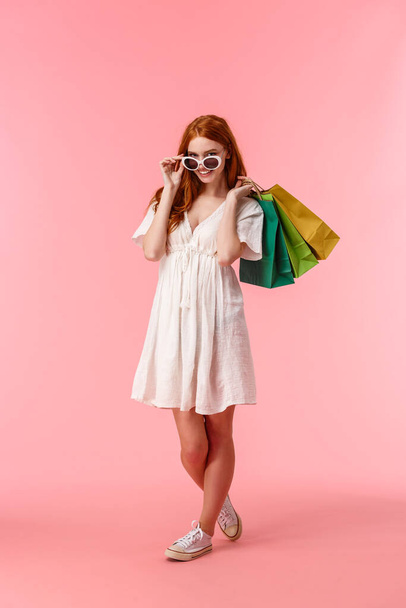 Full-length vertical shot alluring happy redhead female release stress with shopping, shopaholic walking along stores, shop-malls with bags behind back, take-off glasses, smiling camera. - Foto, afbeelding