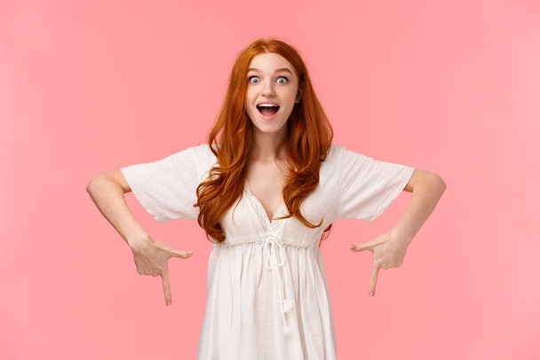 Excited and happy, amazed cheerful redhead woman seing her favorite product in store, pointing fingers down looking with temptation and desire, eager buy it, pink background. - Φωτογραφία, εικόνα