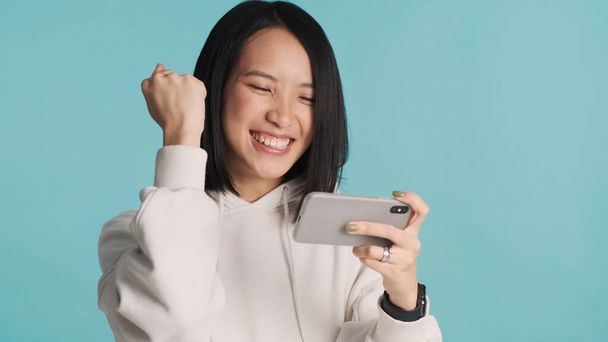 Excited Asian woman emotionally playing on mobile phone celebrating win in online game isolated on blue background. Winner expression - Photo, Image