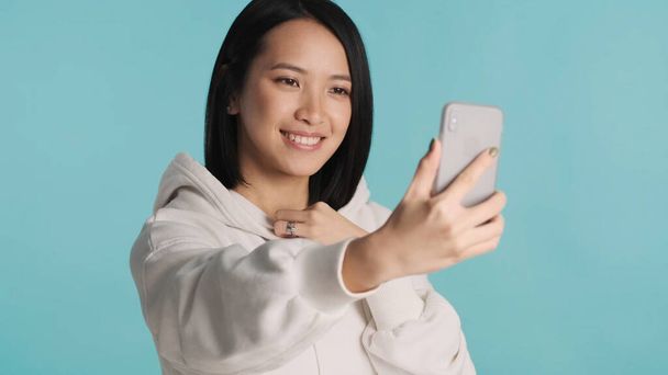 Азиатская женщина выглядит мило искренне улыбаясь, делая селфи на смартфоне на синем фоне - Фото, изображение